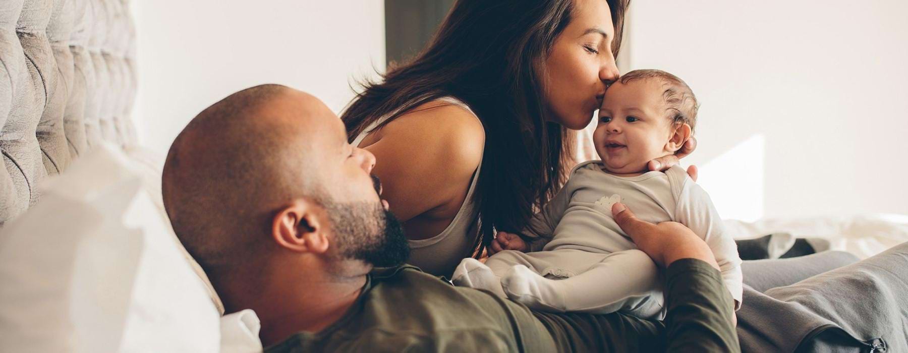 mother kisses her infant while father lies in bed and holds him