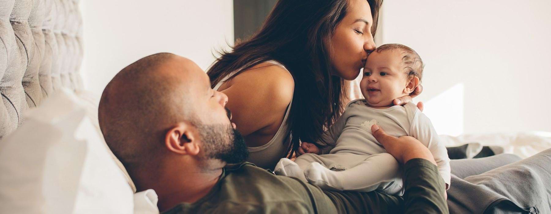 mother kisses her infant while father lies in bed and holds him